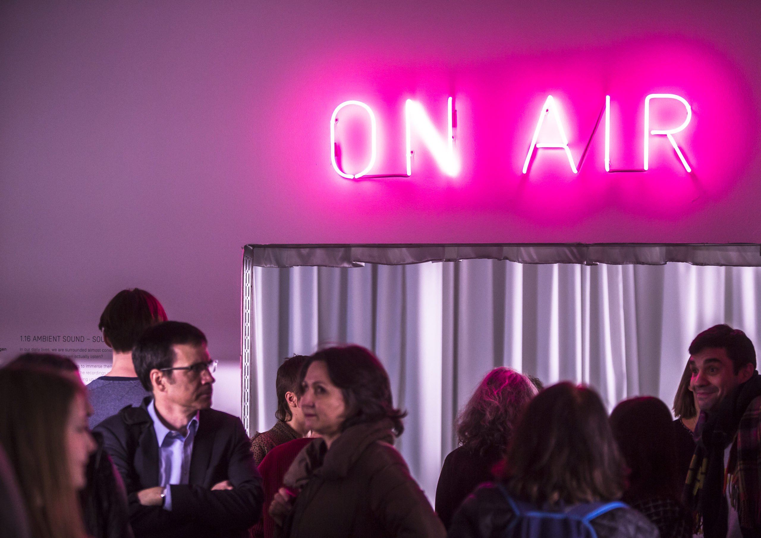A pink "ON AIR" neon sign glows above a group of people in a modern room.
