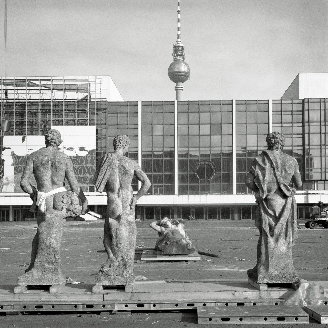 Three ancient statues stand in front of a modern building with the Berlin TV Tower in the background. Black-and-white photo.