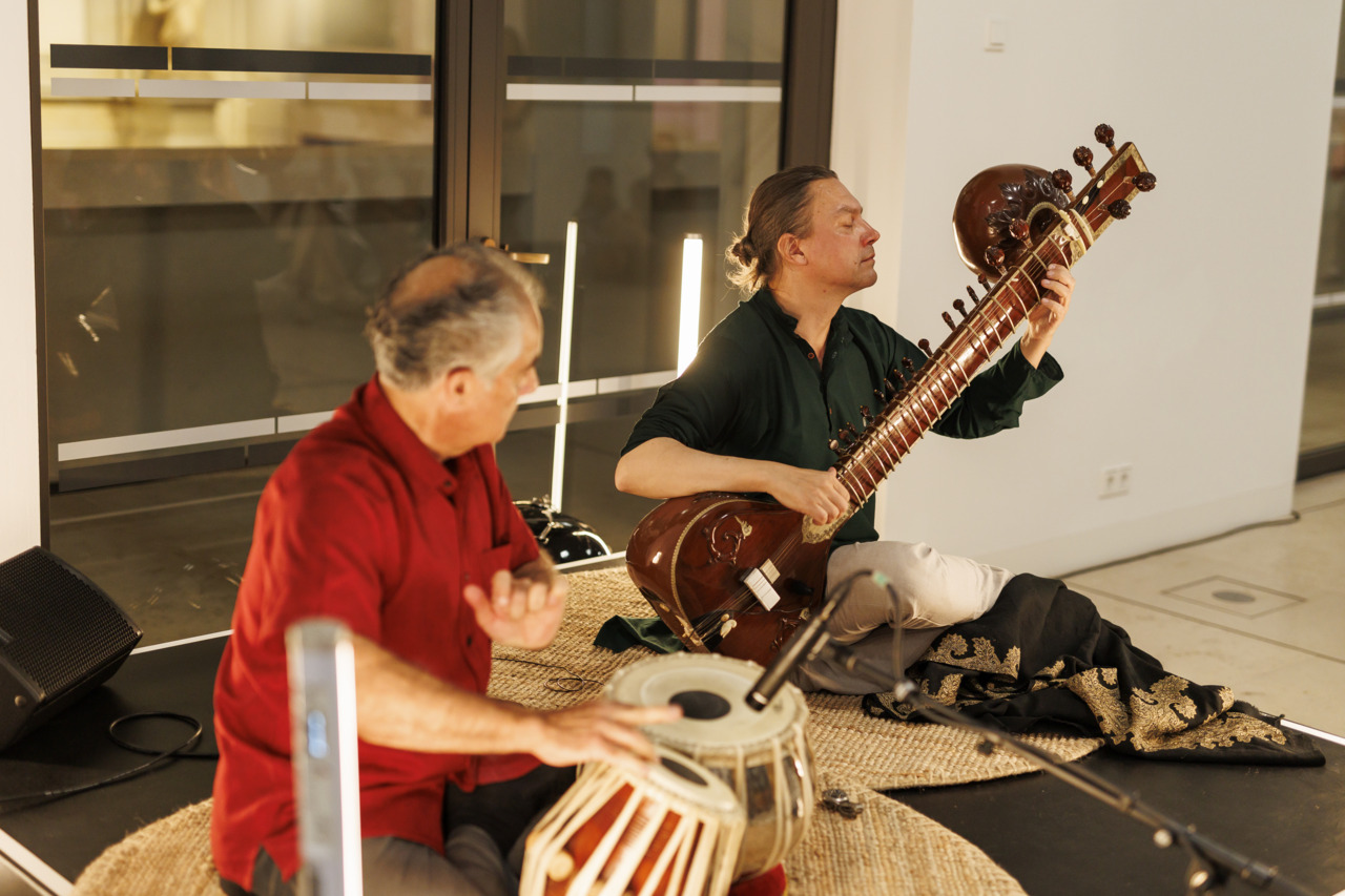 Zwei Musiker spielen live in einem modernen Raum: ein Mann auf einer Sitar und ein anderer auf einer indischen Trommel.