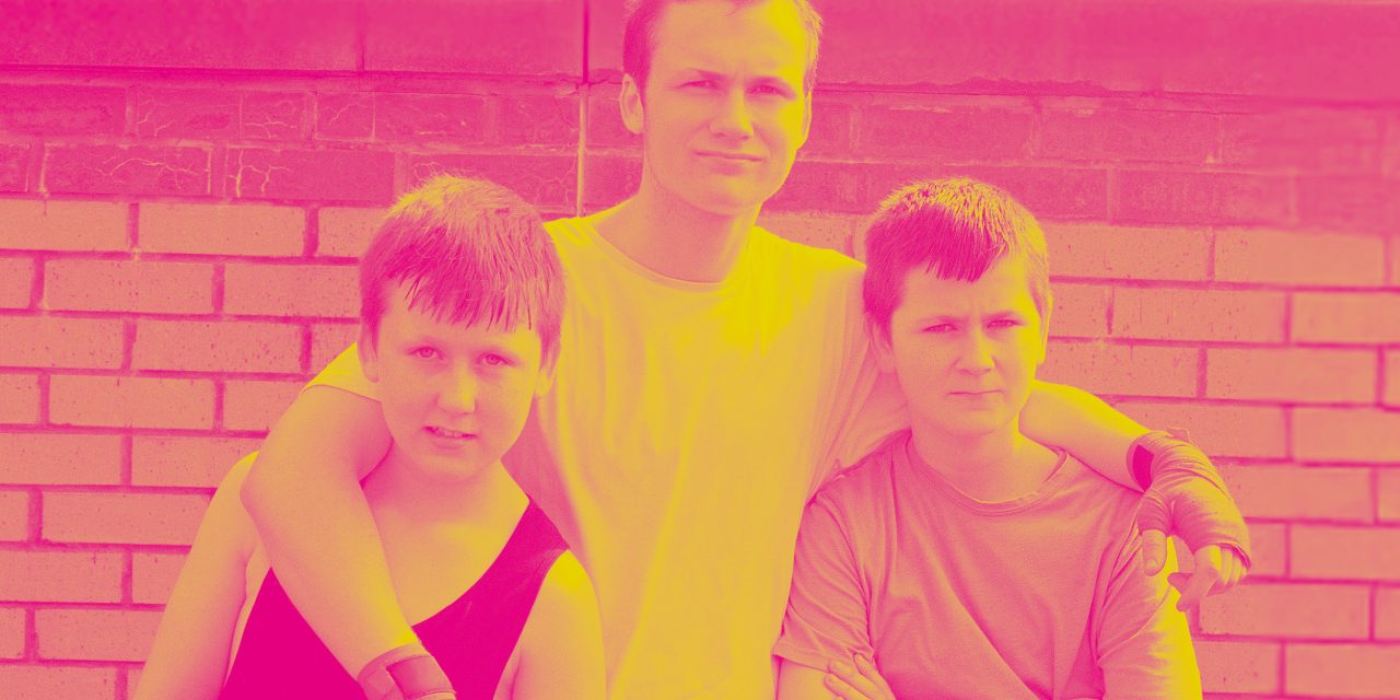 Three teenagers pose in front of a brick wall, one has his arm around another, and the colors are vibrant.