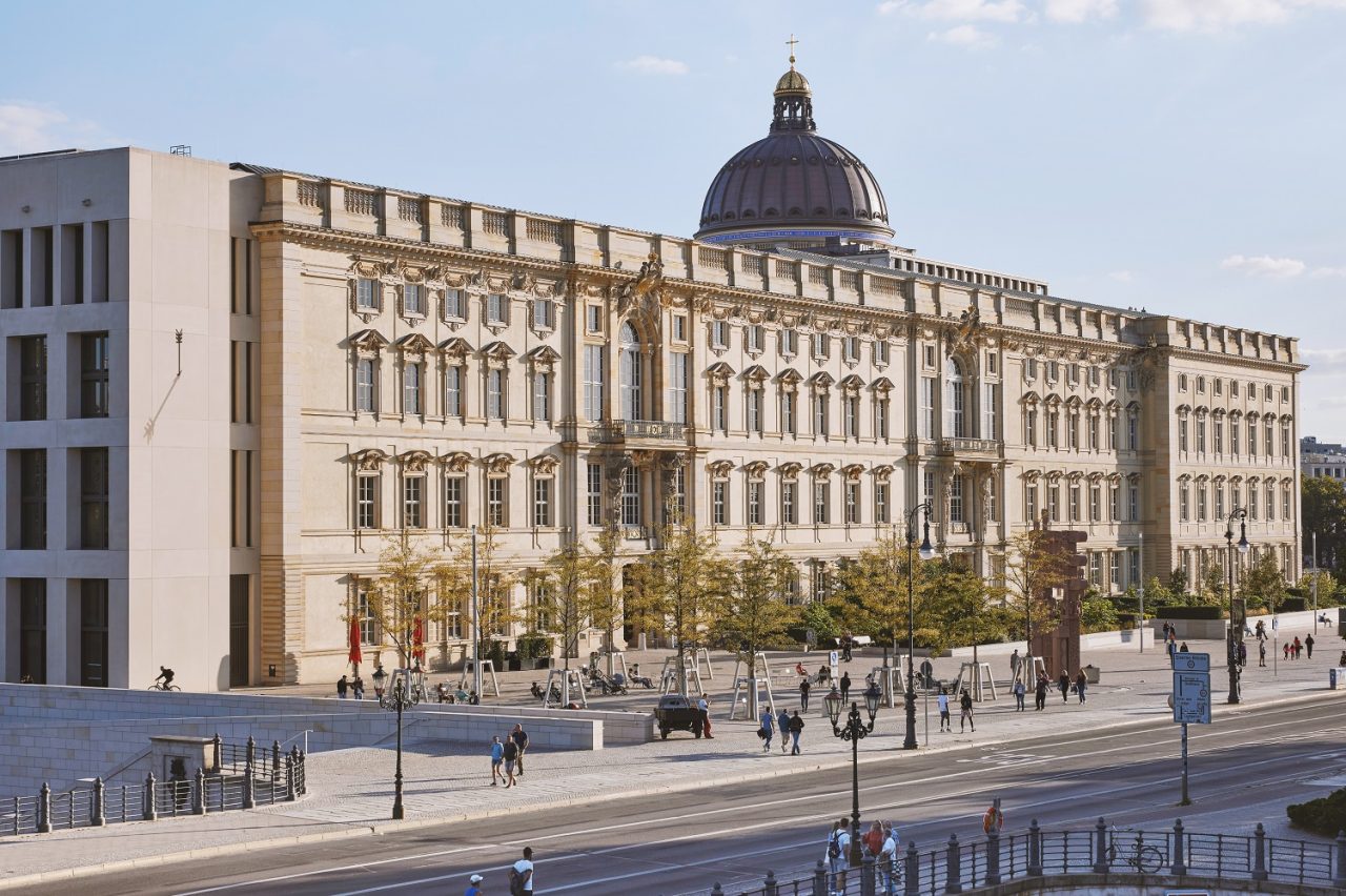 Historic building with a dome and modern architecture, surrounded by people and trees.