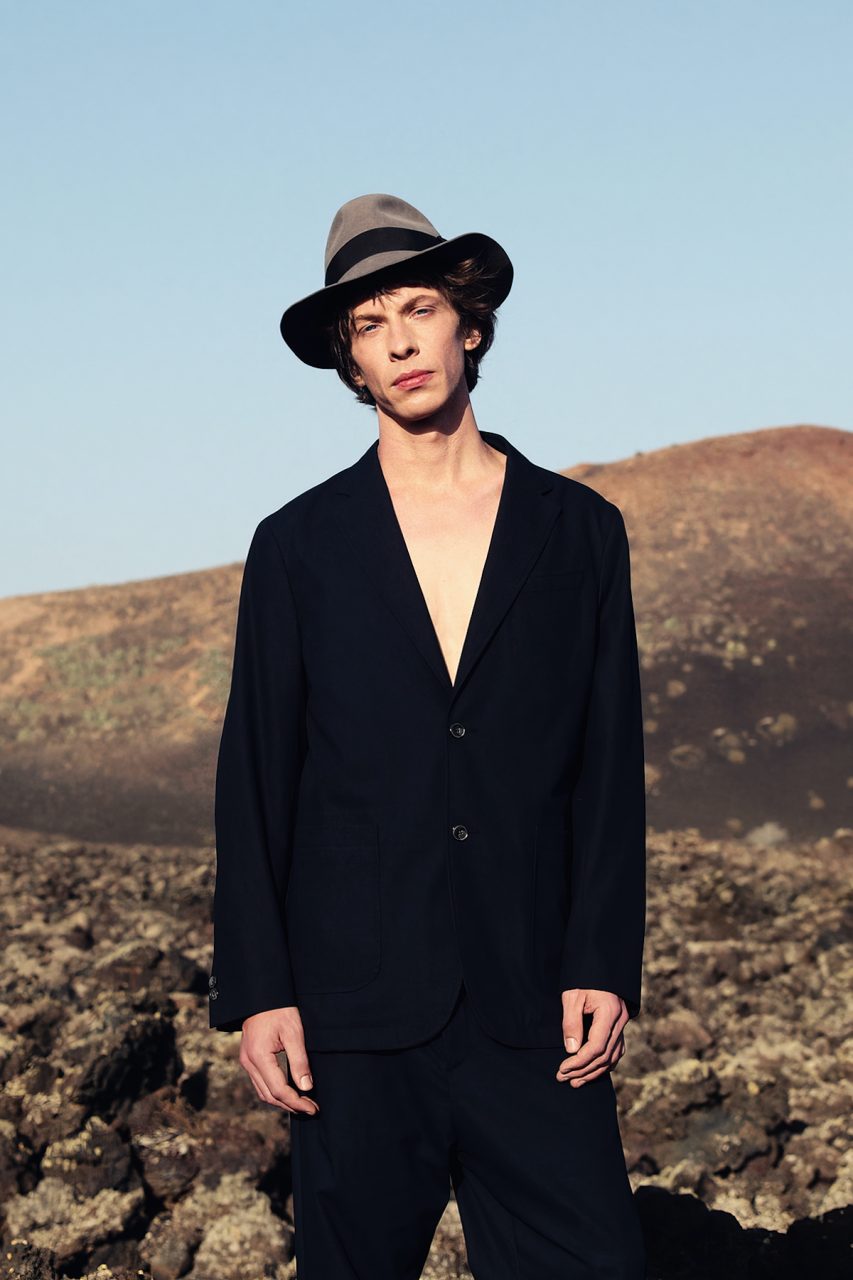 Modischer Mann in schwarzer Jacke und Hut steht vor vulkanischer Landschaft unter klarem, blauem Himmel.