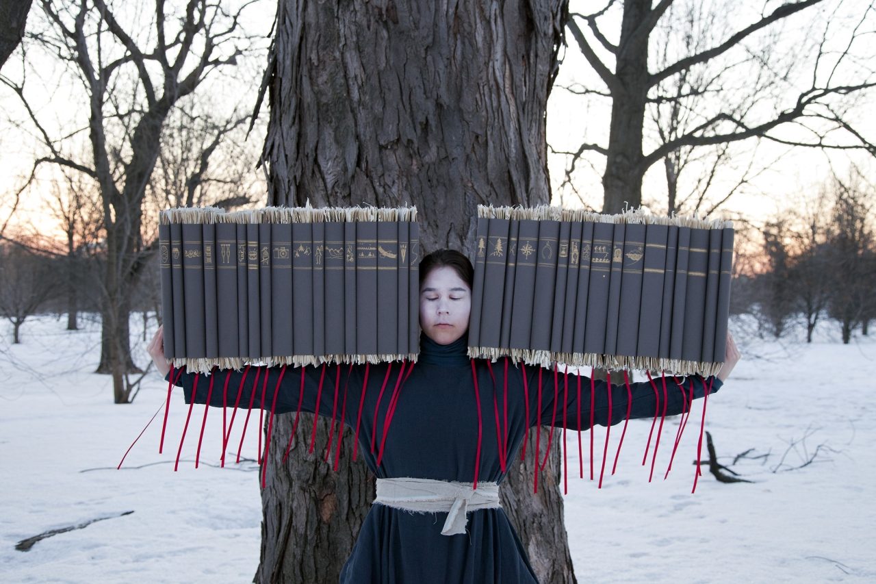 A girl in nature carries books with red ribbons in front of a tree in the snow as the sun sets.
