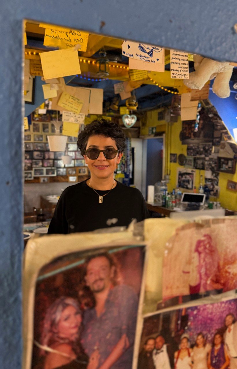 Frau mit kurzen Haaren und Sonnenbrille steht in einem bunten Raum, dekoriert mit Fotos und Notizen.