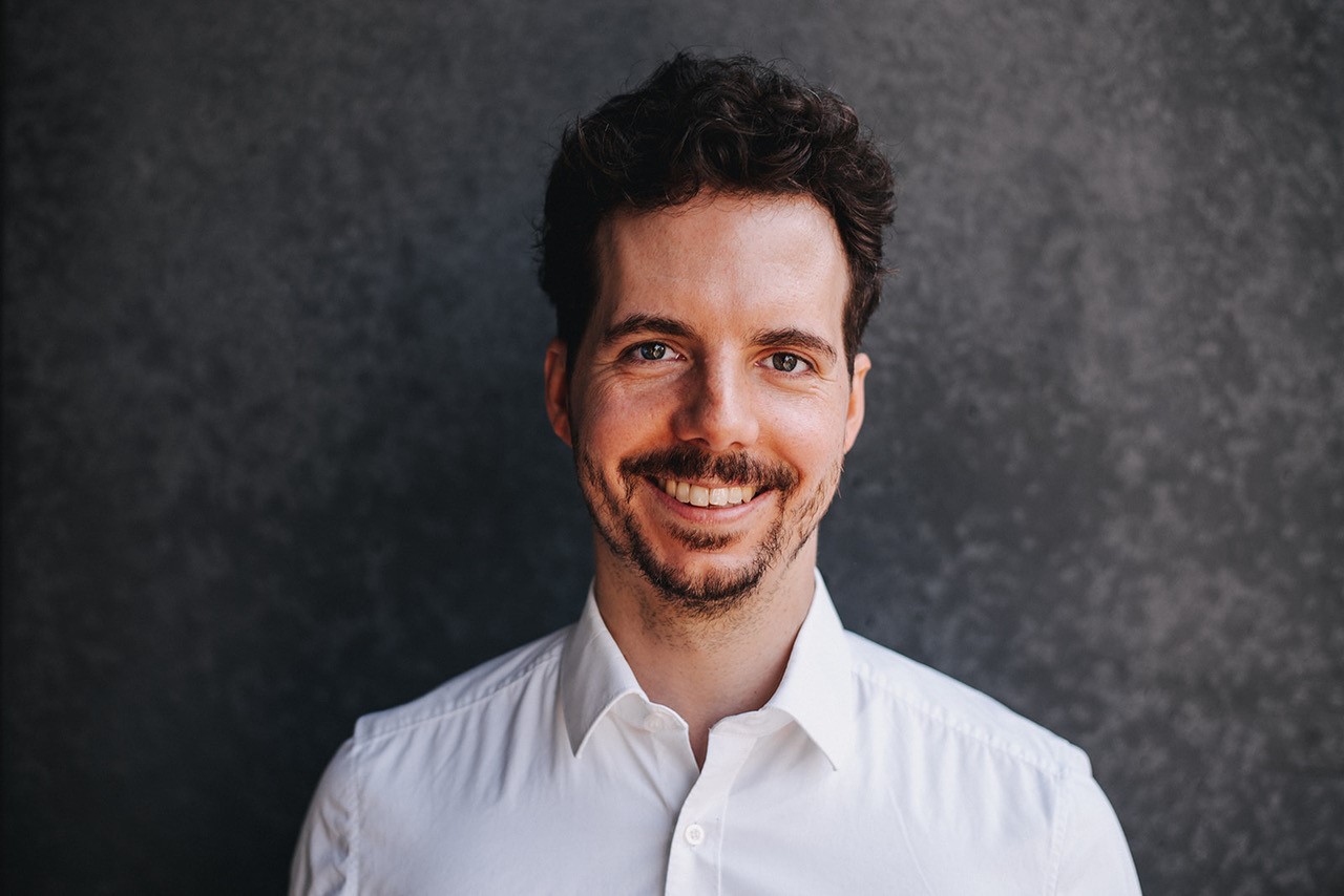 Smiling man with short hair, wearing a white shirt against a dark background.