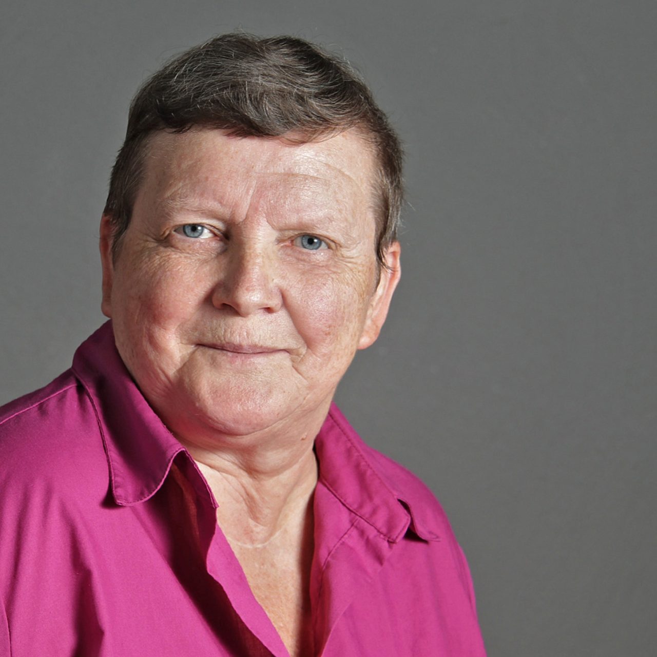 Portrait of an older woman with short gray hair in a pink shirt, smiling against a gray background.