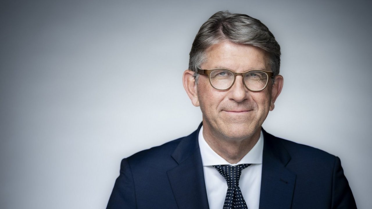 Portrait of a man in glasses and a suit, smiling against a neutral background.