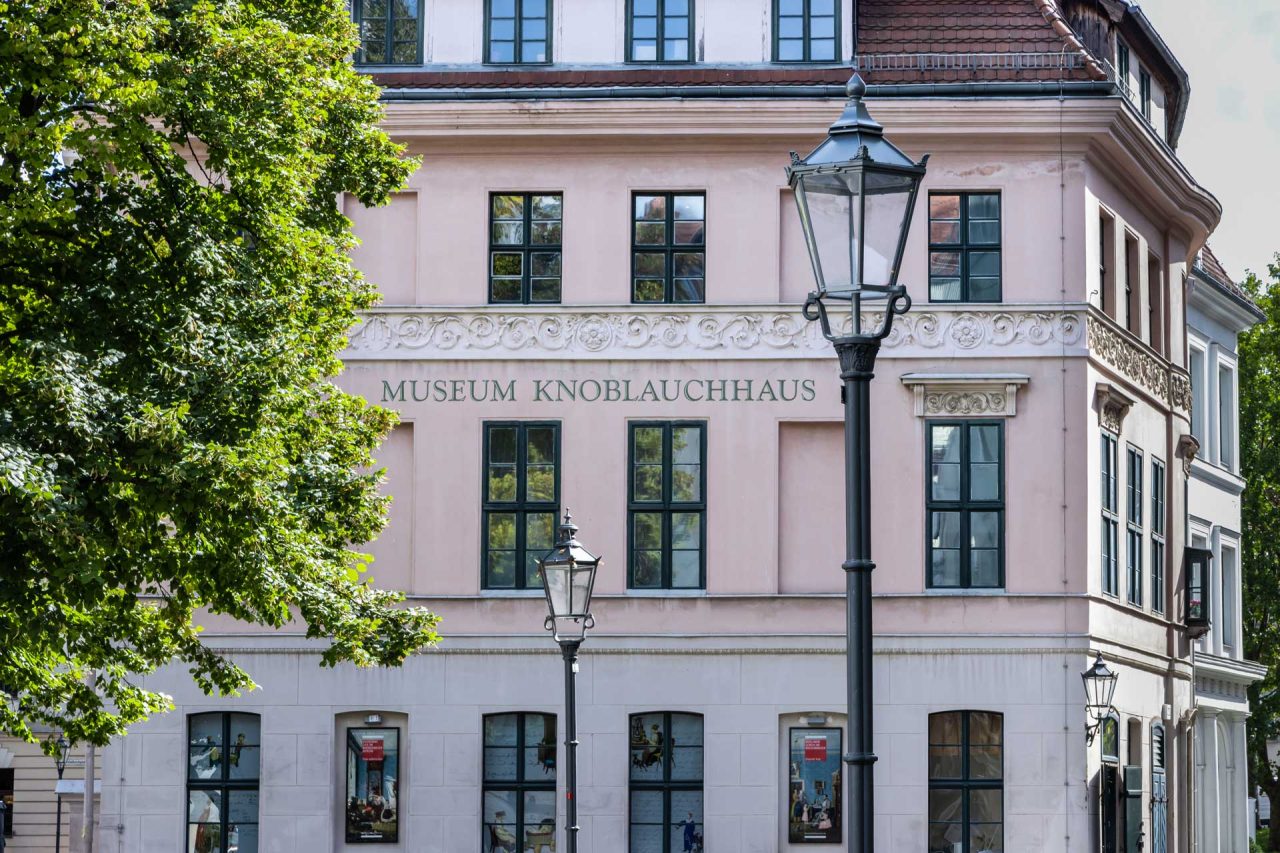 Fassade des Museums Knoblauchhaus mit Fenstern und Straßenlaternen, umgeben von Bäumen.