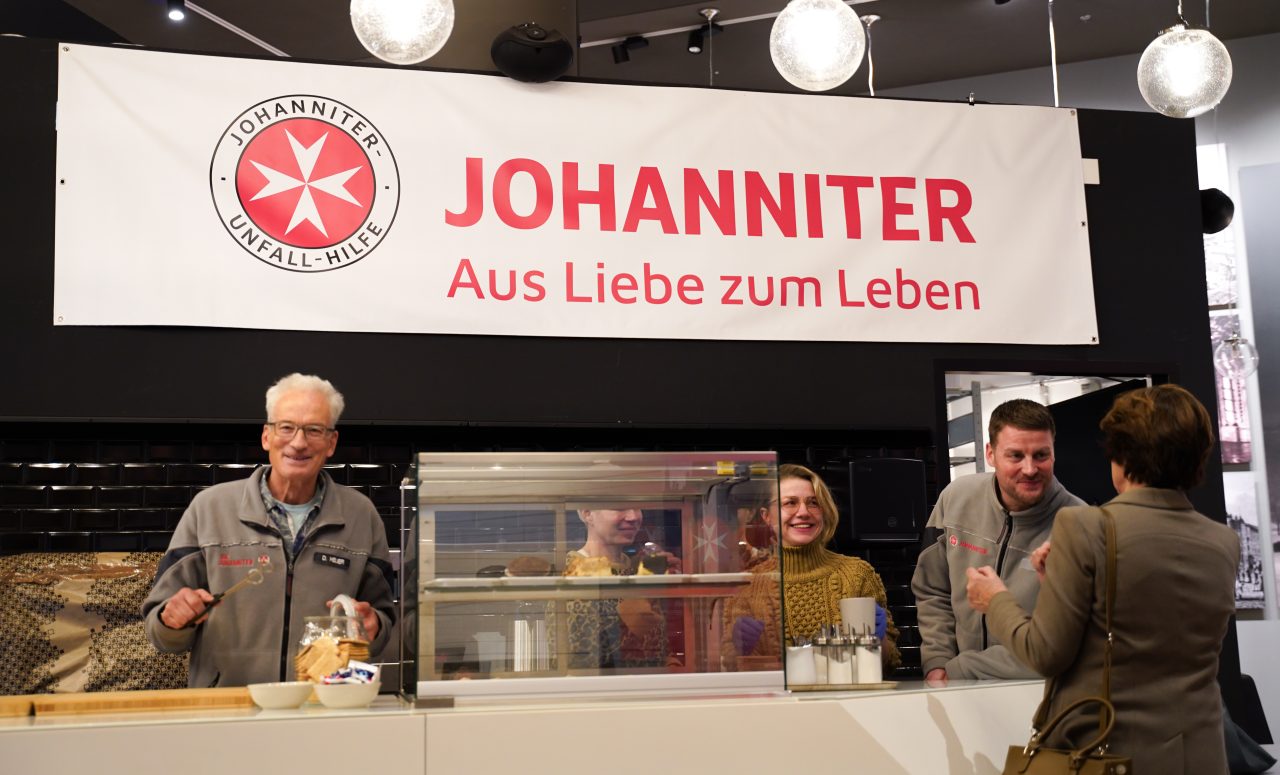 People at the Johanniter booth speaking friendly, offering coffee and snacks. Banner in the background.