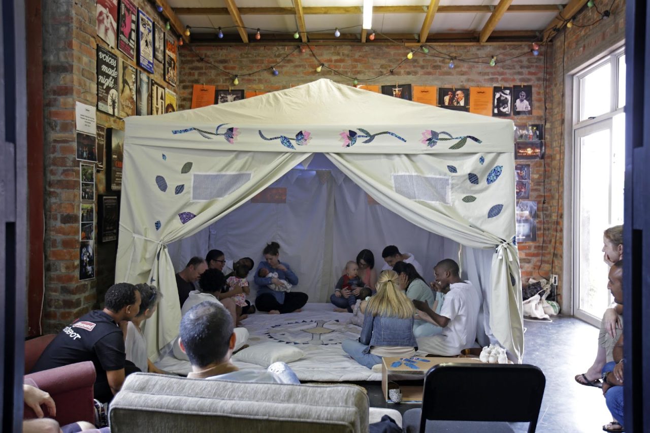 Gruppe von Menschen in einem Zelt, die gemeinsam sitzen und interagieren, umgeben von bunten Postern und Lichtern.