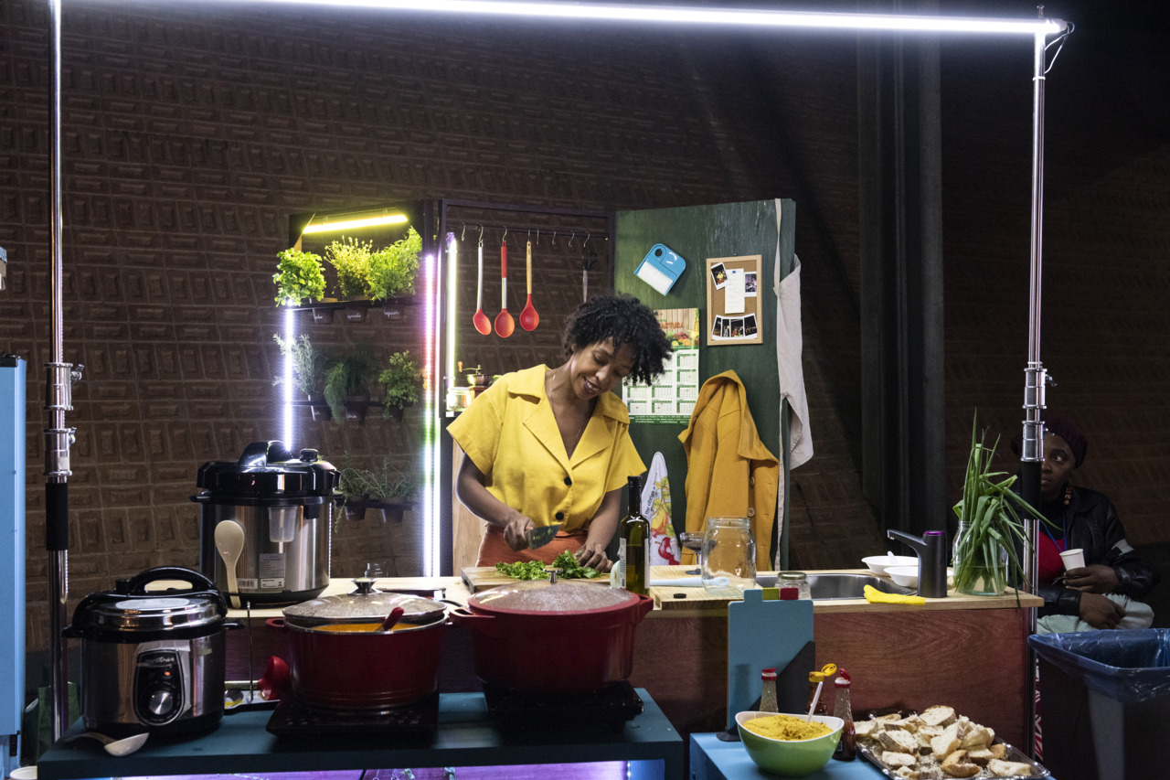 A woman in a yellow outfit is cutting vegetables in a modern kitchen with colorful accessories.