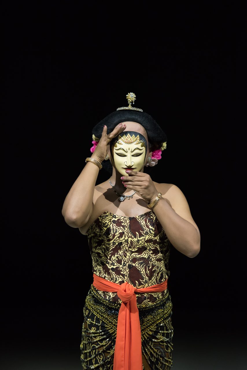 Woman in traditional clothing holds a mask, ready for a performance against a black background.