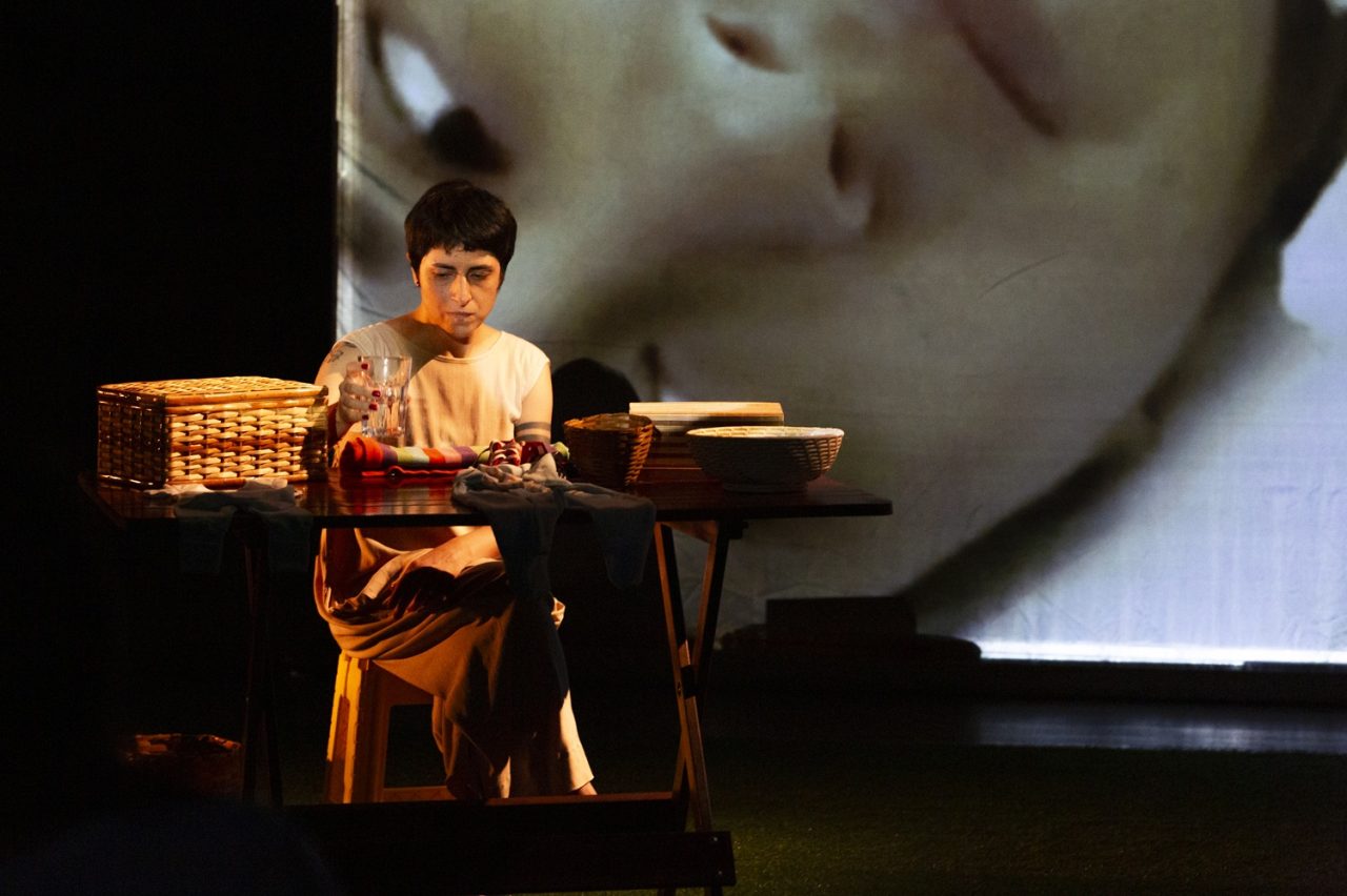 A person sits at a table with laundry and a glass, with a large picture visible in the background.