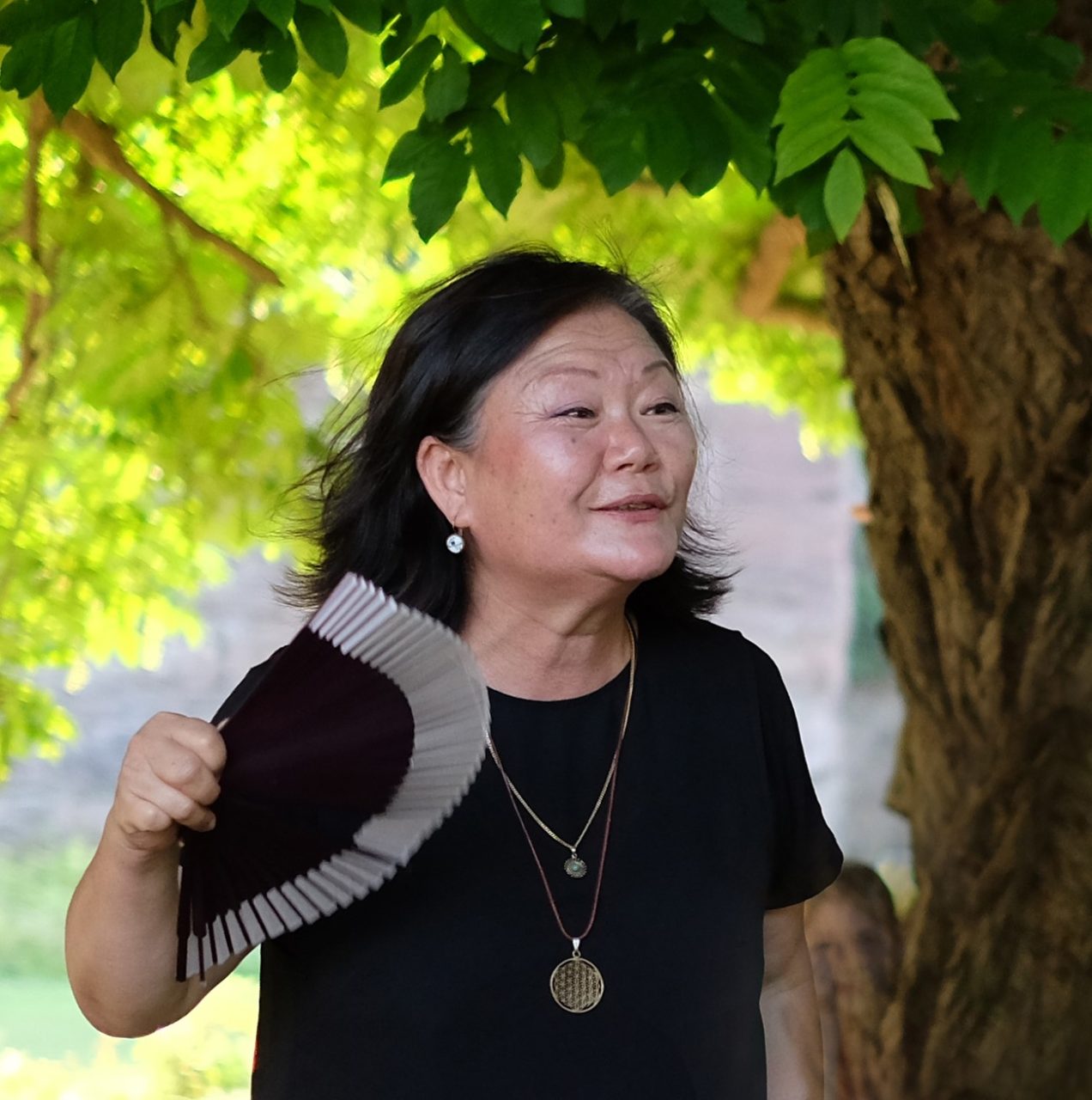 A woman with a fan stands under a tree, laughing while talking to someone.