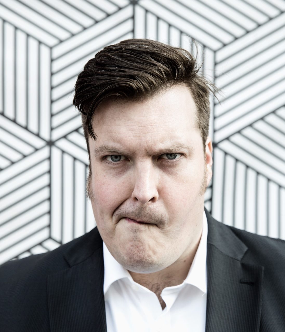 Man in a suit with a serious expression in front of a geometric background.