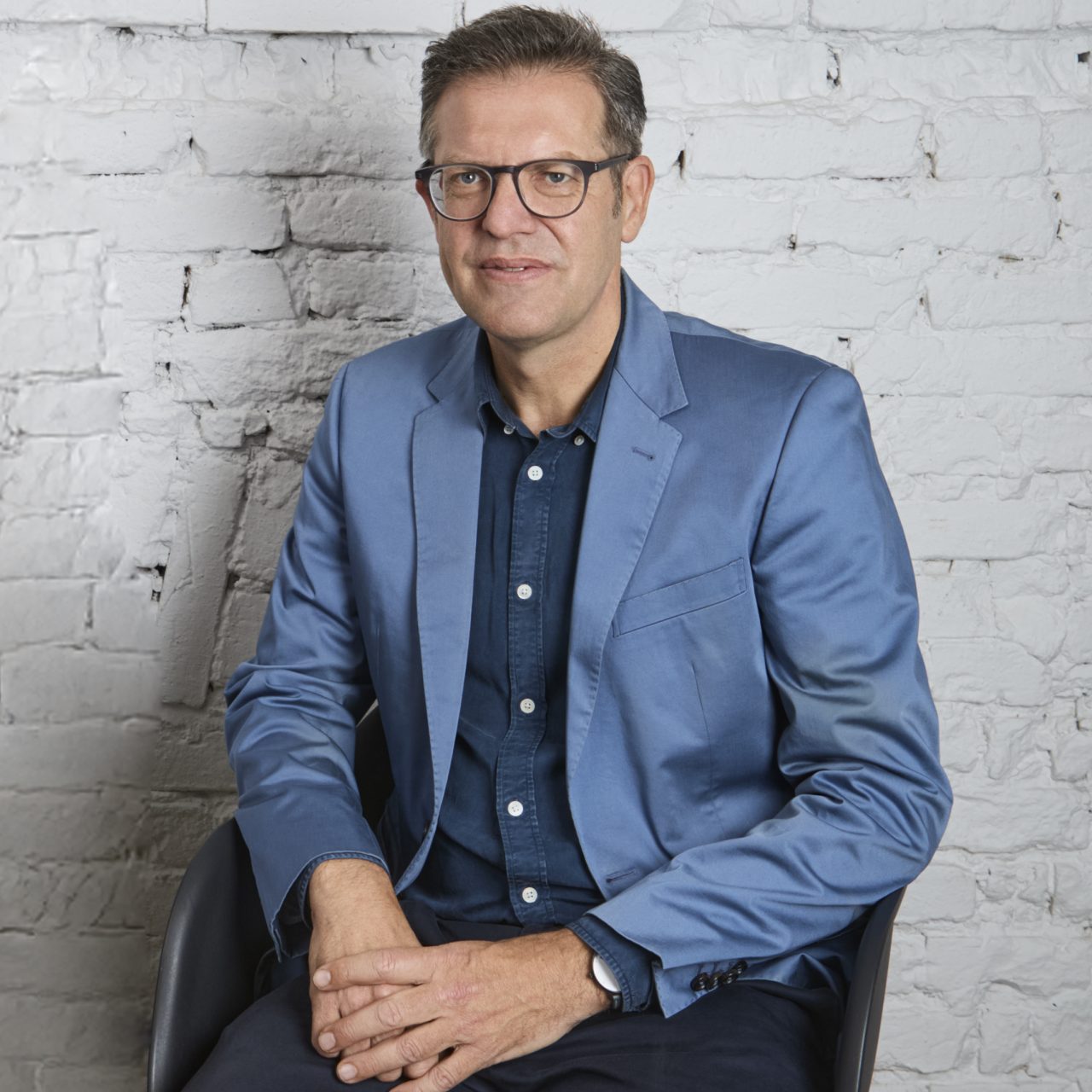 Man in a blue blazer sits in front of a white brick wall and smiles slightly.