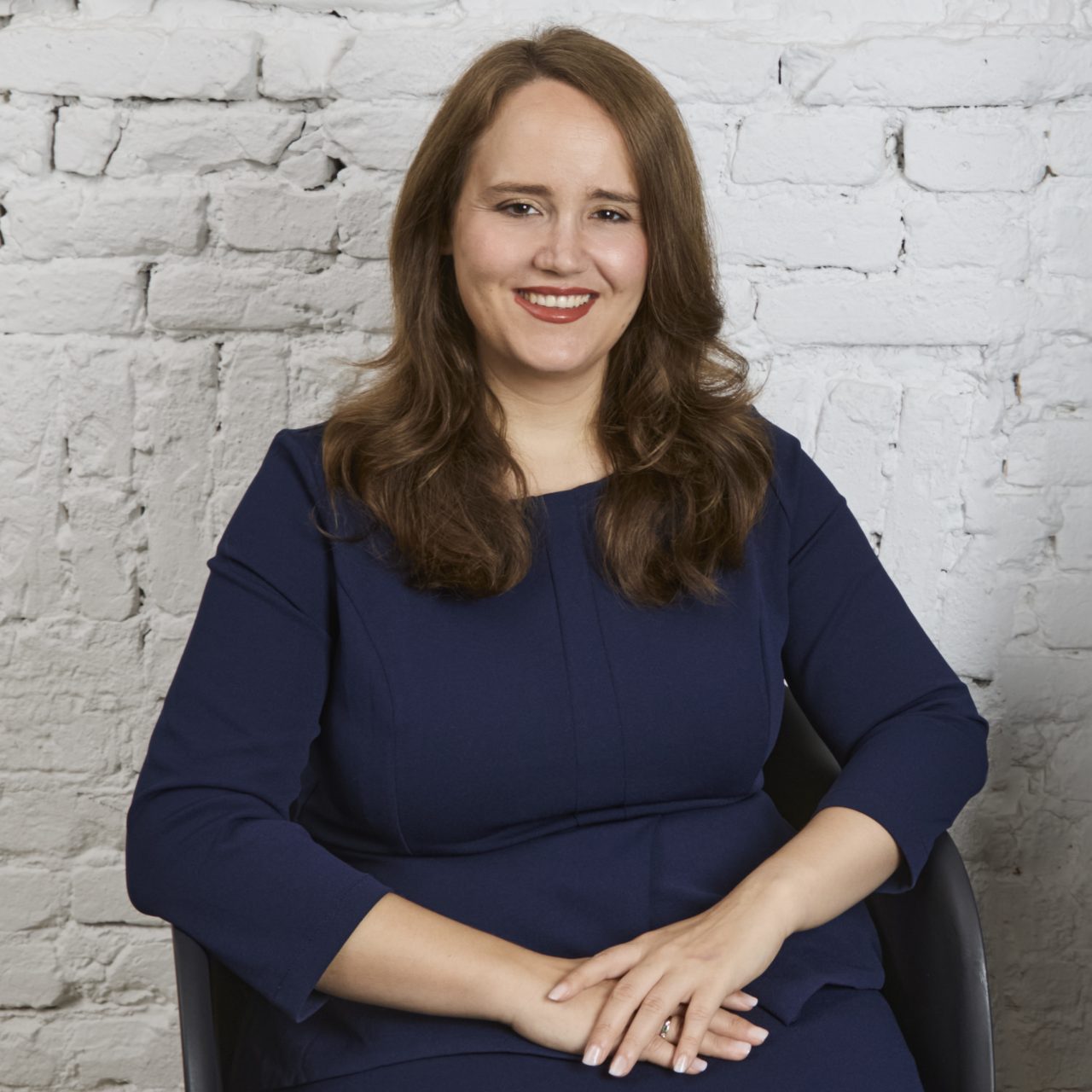 Portrait of a smiling woman with long hair in a blue dress, sitting in front of a white brick wall.