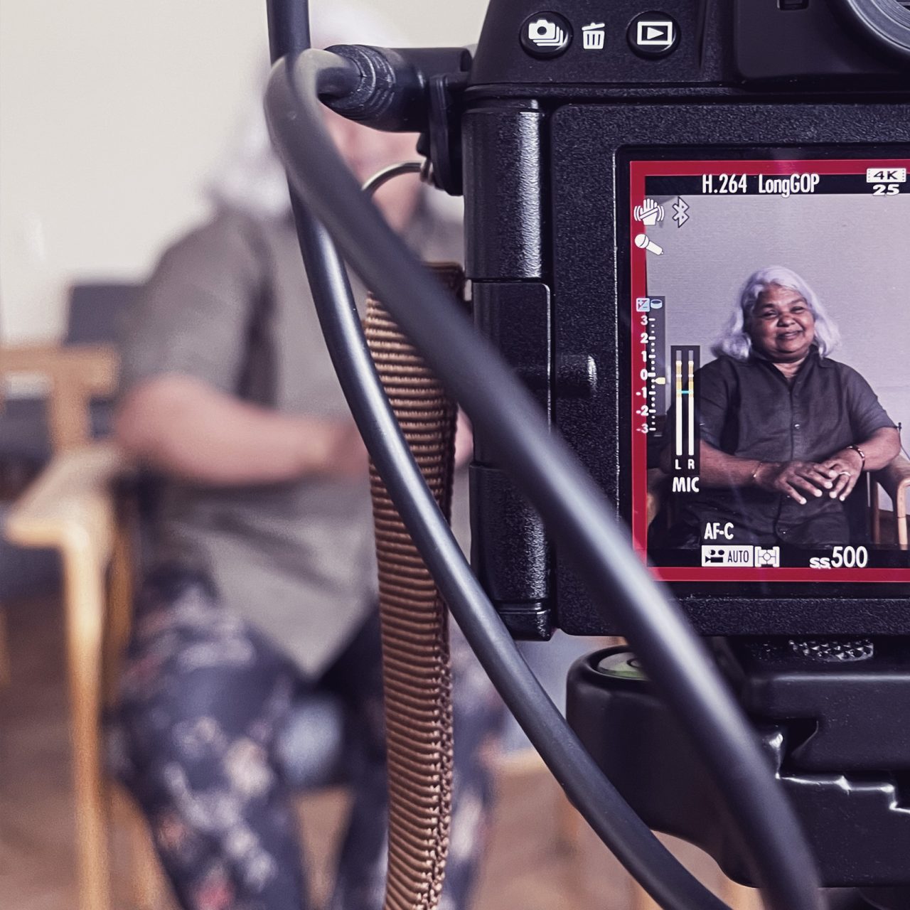 Kameraaufnahme einer Person mit grauen Haaren im Fokus, während eine zweite Person im Hintergrund unscharf bleibt.