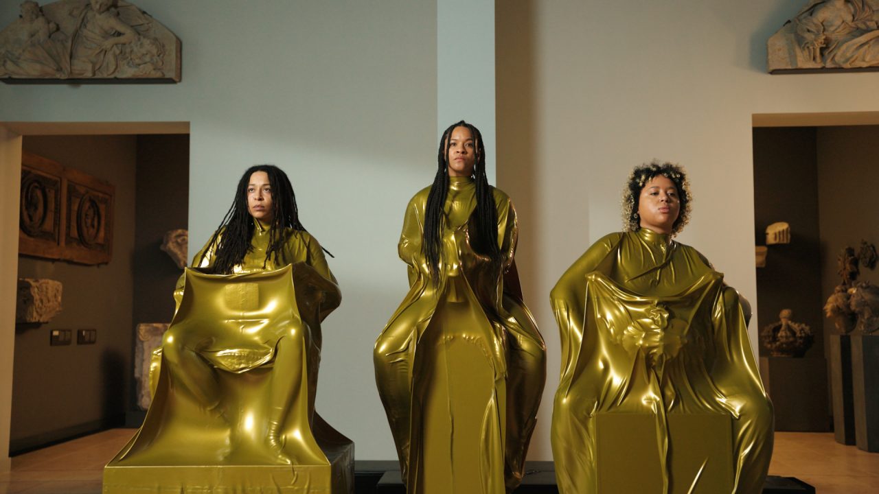 Drei Personen in goldenen, fließenden Gewändern sitzen in einer Galerie mit antiken Statuen im Hintergrund.