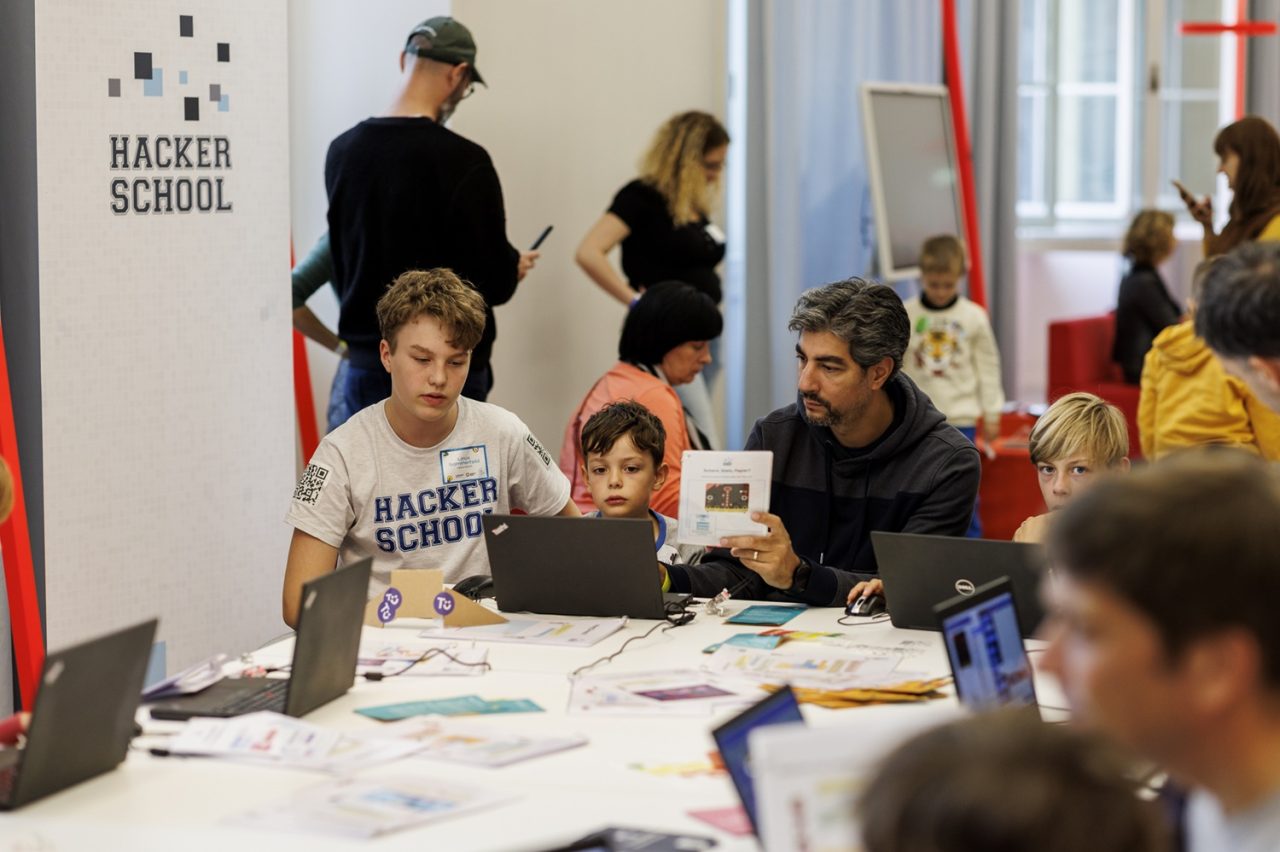 Children and adults are working together on laptops in a learning environment with a "Hacker School" banner in the background.