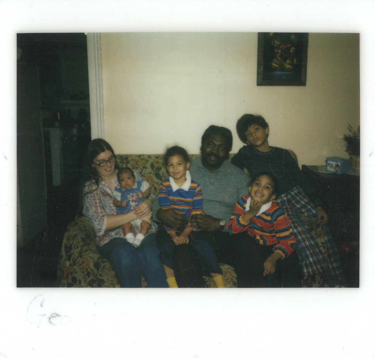 A family sits smiling on a sofa, surrounded by children and a baby, in a cozy room.
