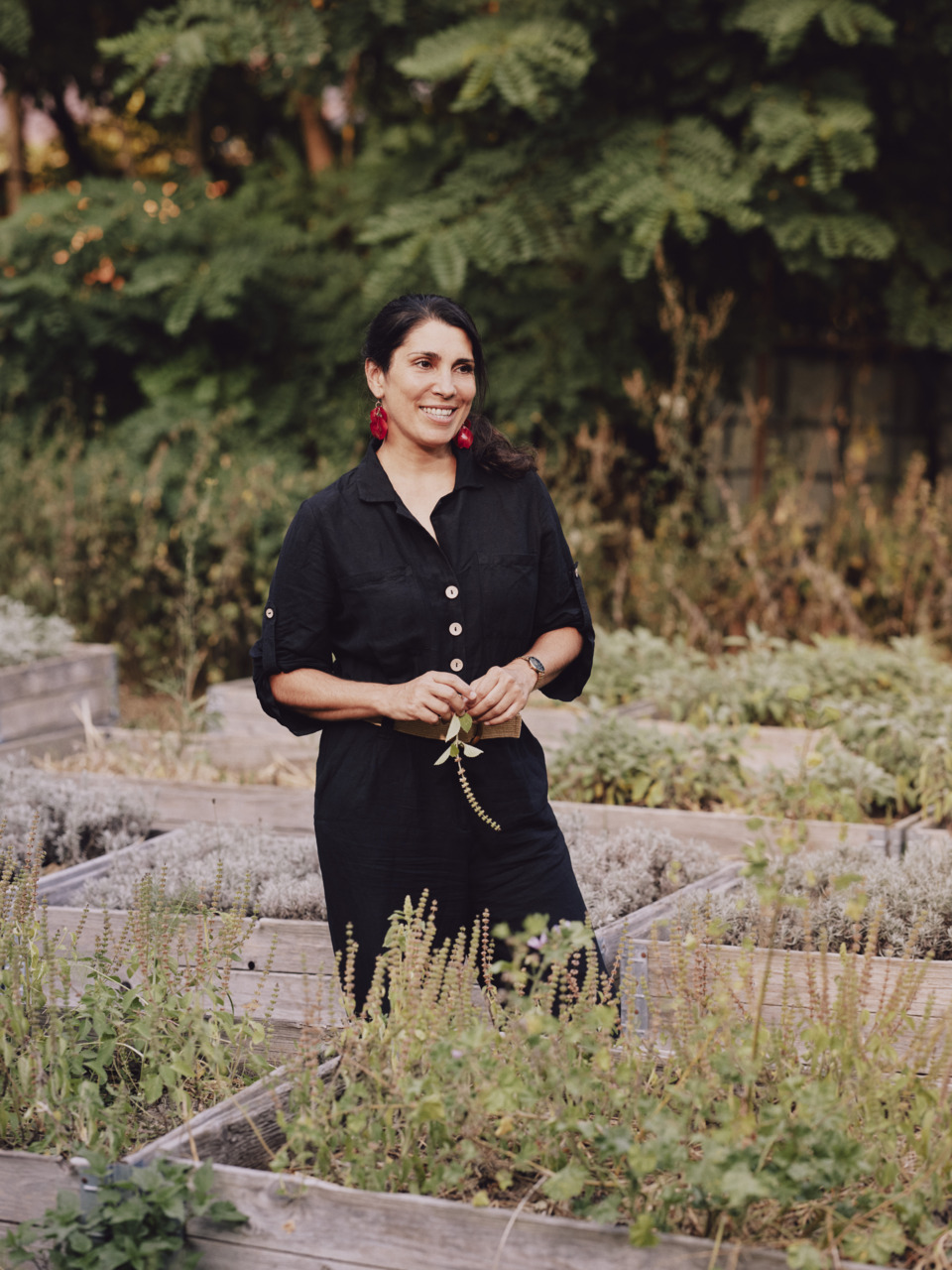 Frau in schwarzem Outfit steht in einem Kräutergarten, umgeben von grünen Pflanzen und Holzbeeten, und lächelt freundlich.