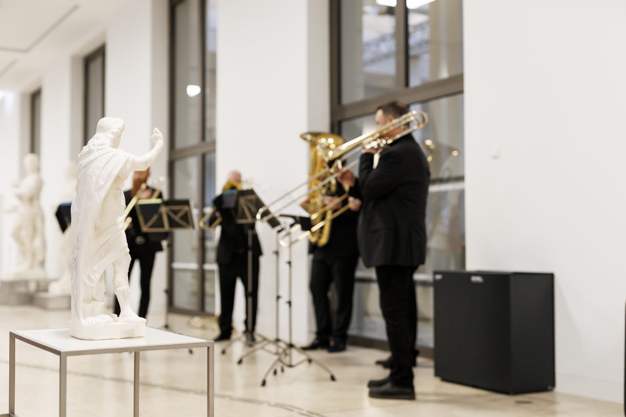 Musicians with trombones play in a modern exhibition space, while a statue stands in the foreground.