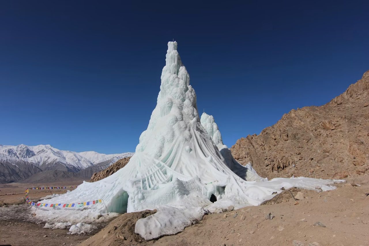 Icy statue in a mountainous landscape, surrounded by snow and colorful prayer flags under a clear blue sky.