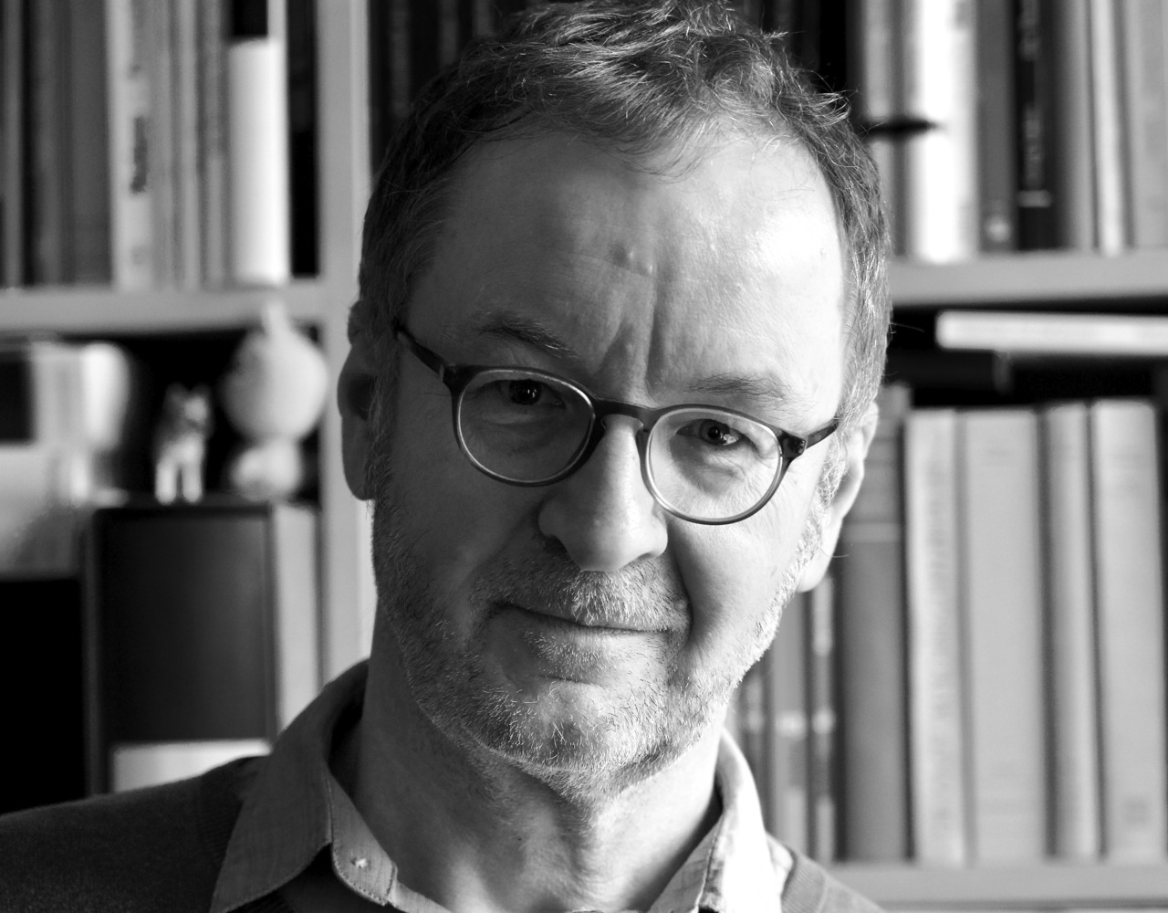 Porträt eines lächelnden Mannes mit Brille vor einem Bücherregal in Schwarzweiß.