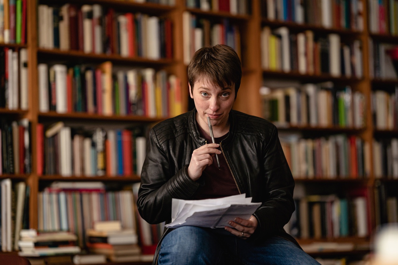 Eine Person in schwarzer Jacke sitzt in einer Bibliothek, hält einen Stift und schaut nachdenklich auf Manuskriptblätter.