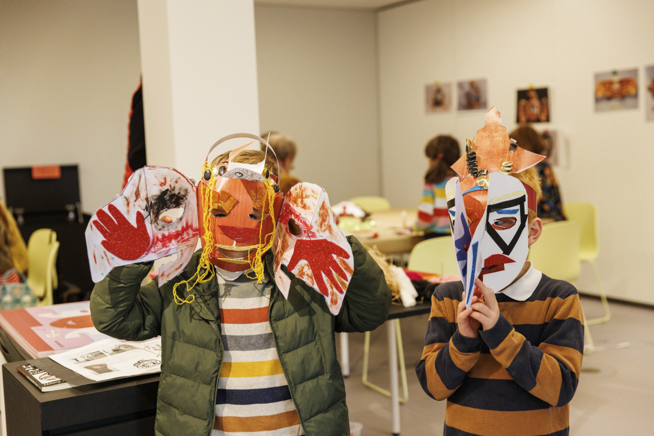 Zwei Kinder tragen bunte, selbstgemachte Masken in einem Kreativraum mit anderen Kindern und Bastelmaterialien.