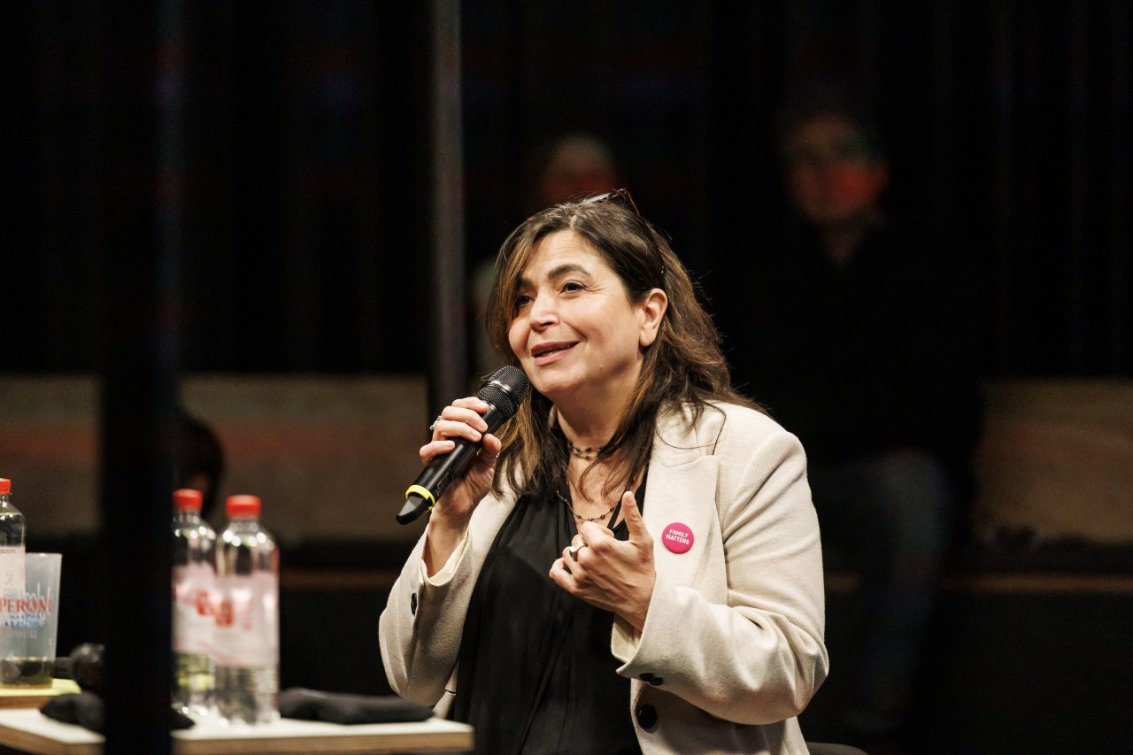 Frau mit Mikrofon spricht leidenschaftlich auf einer Veranstaltung, im Hintergrund sind Wasserflaschen zu sehen.