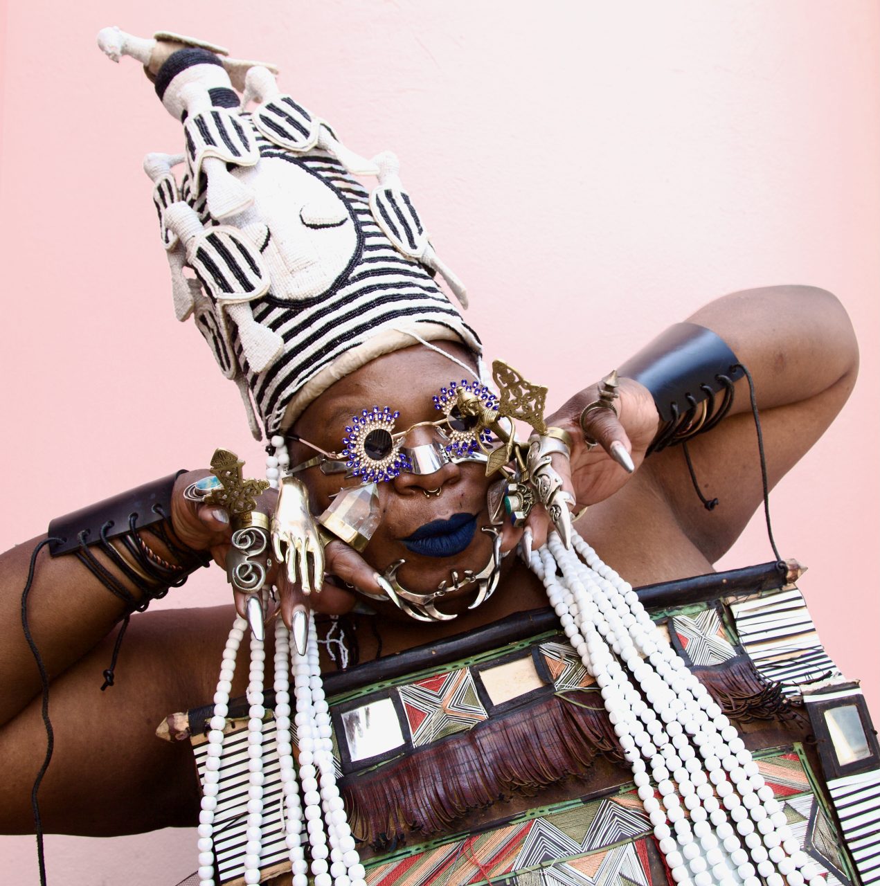 Kreative Person in ikonischer Kopfbedeckung und auffälligem Schmuck posiert vor einer rosa Wand.