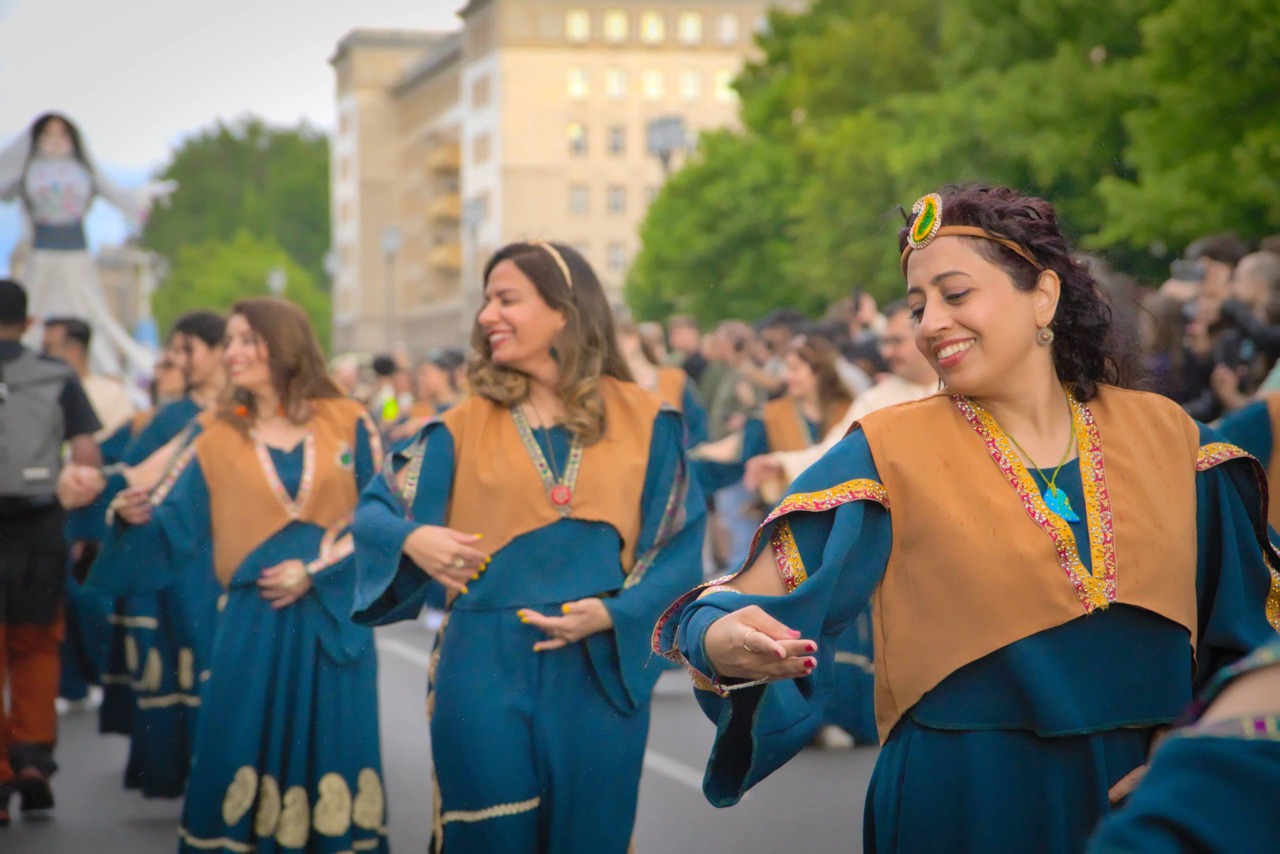 Frauen in traditionellen Kleidern tanzen fröhlich bei einem Festumzug. Im Hintergrund sind Publikum und grüne Umgebung.