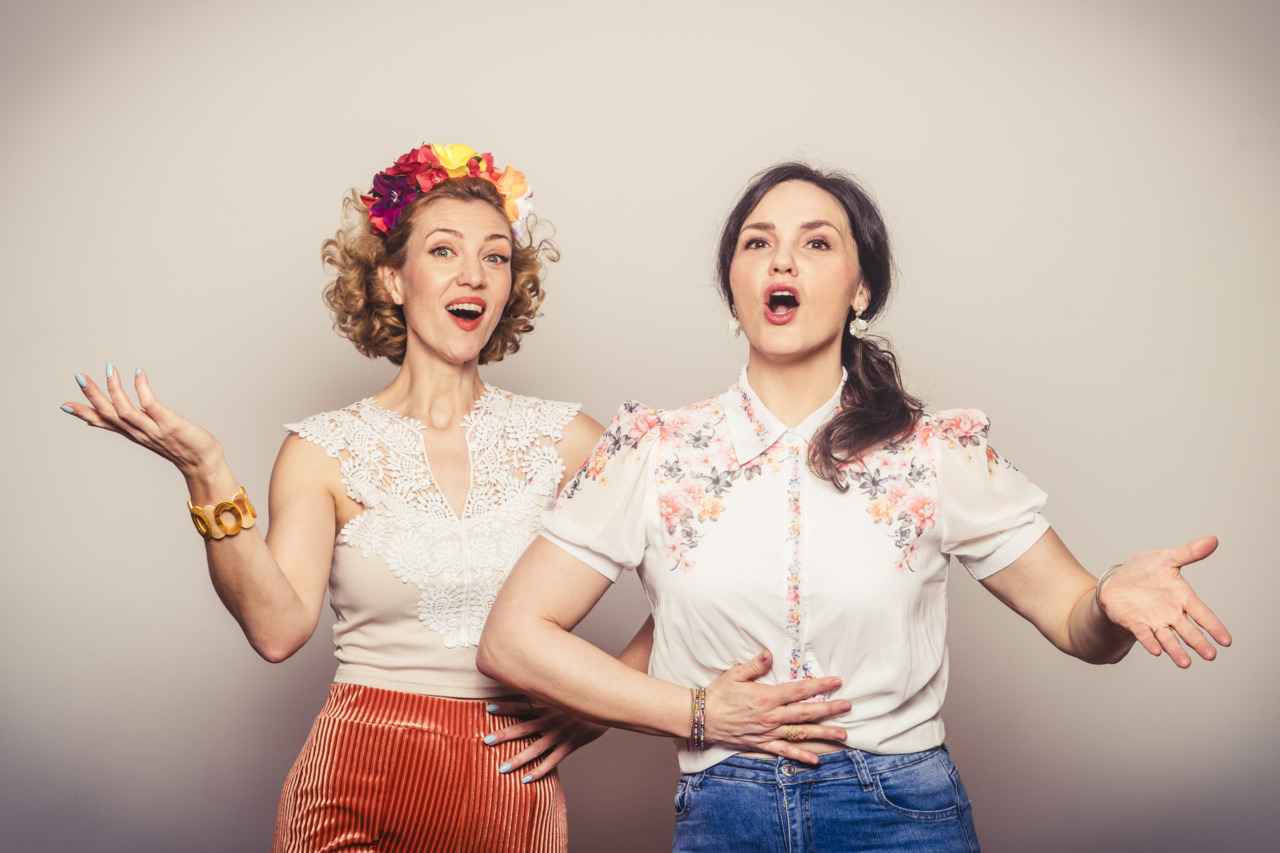 Two women sing joyfully, one with a flower crown, the other in a floral blouse, both radiating enthusiasm.