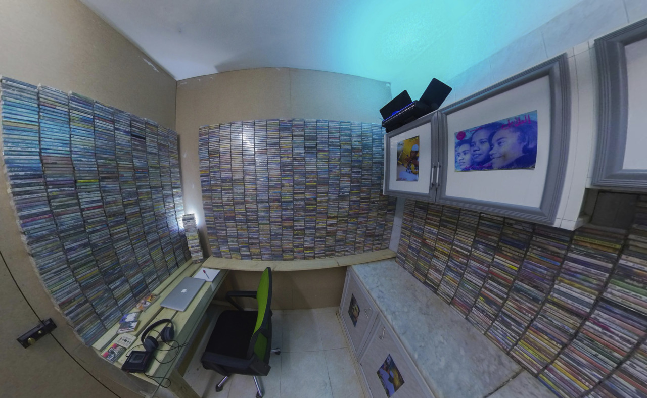 Kleinraum mit Wänden aus Kassettensammlungen, Schreibtisch, Laptop und Fotos im sanften Licht.