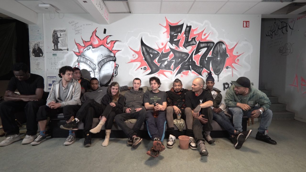 Gruppe von Menschen auf einem Sofa vor einer mit Graffiti geschmückten Wand, lächelnd und entspannt.