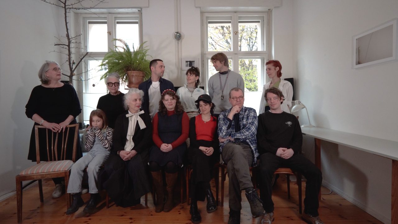 Eine Gruppe von Personen sitzt in einem Raum mit Fenstern und Pflanzen, lächelt und trägt verschiedene Outfits.