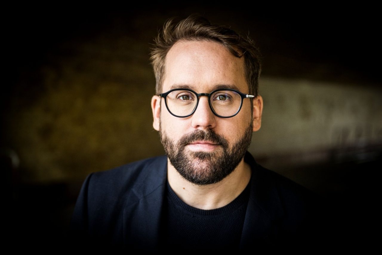 Portrait of a man with glasses and a beard, looking directly at the camera against a dark background.