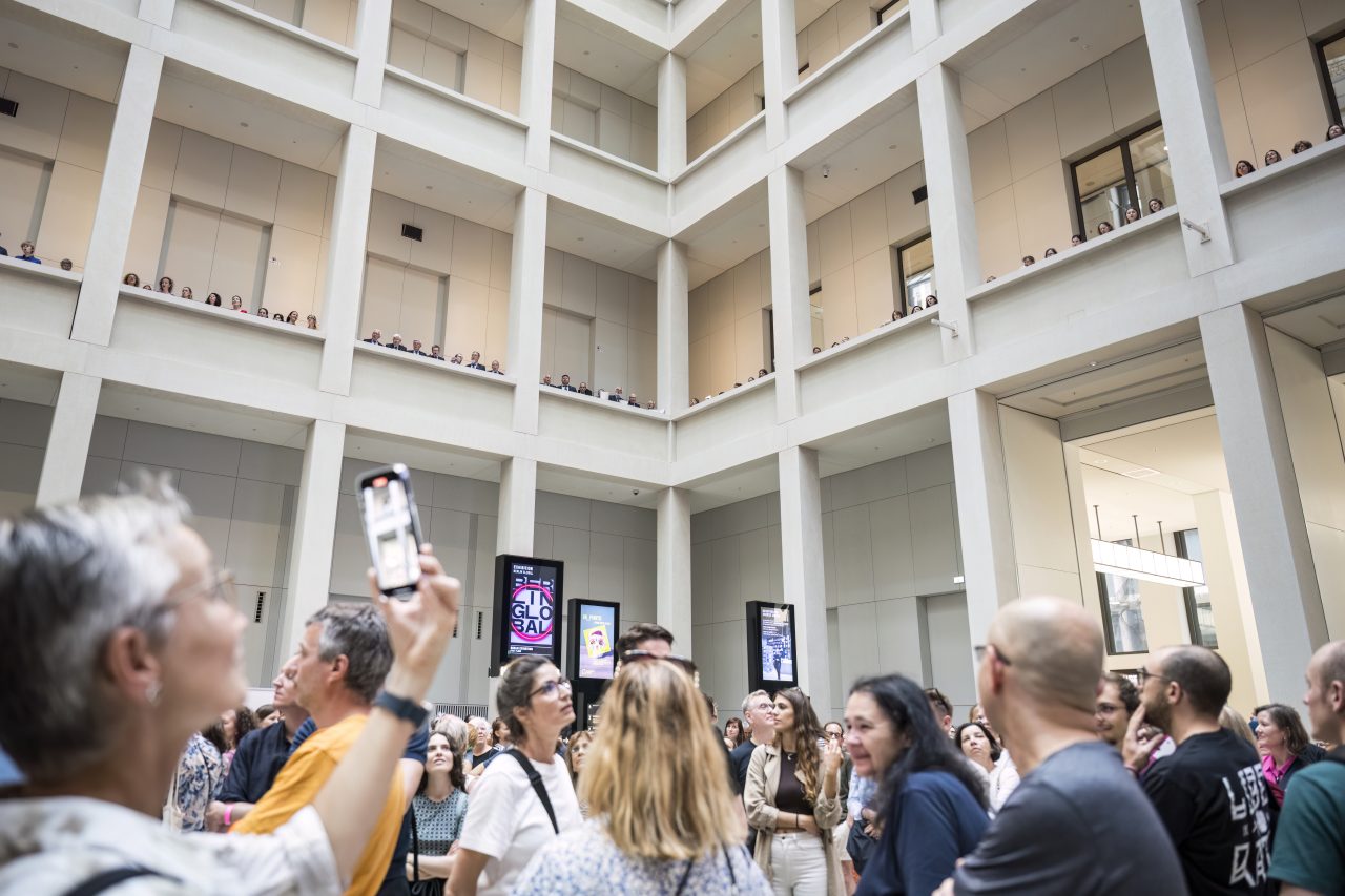 A crowd gathers in a spacious atrium, with people on balconies above observing and taking pictures.