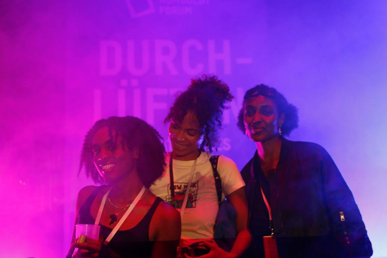Three women smile in colorful light and smoke, in front of a background graphic with the text "AIRING OUT."
