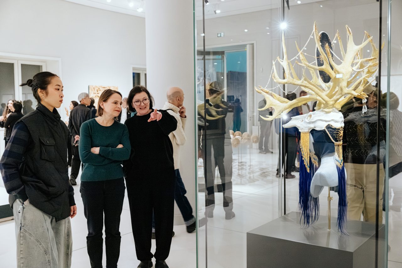 Three people examine an artistic object in a gallery, surrounded by other visitors.