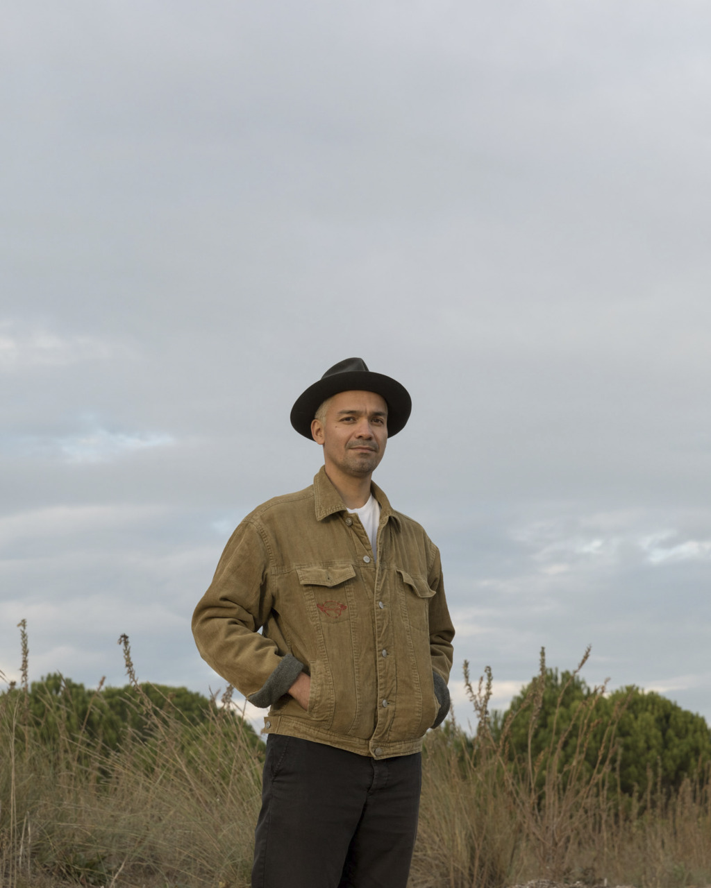 Man in a field wearing a hat and denim jacket, smiling in front of a cloudy sky.