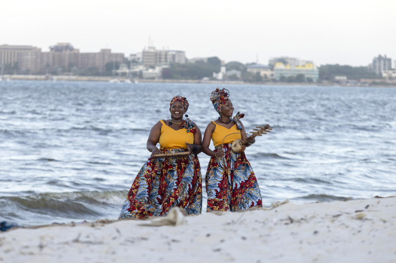 Two women stand on the beach, wearing colorful clothes and smiling while holding musical instruments.