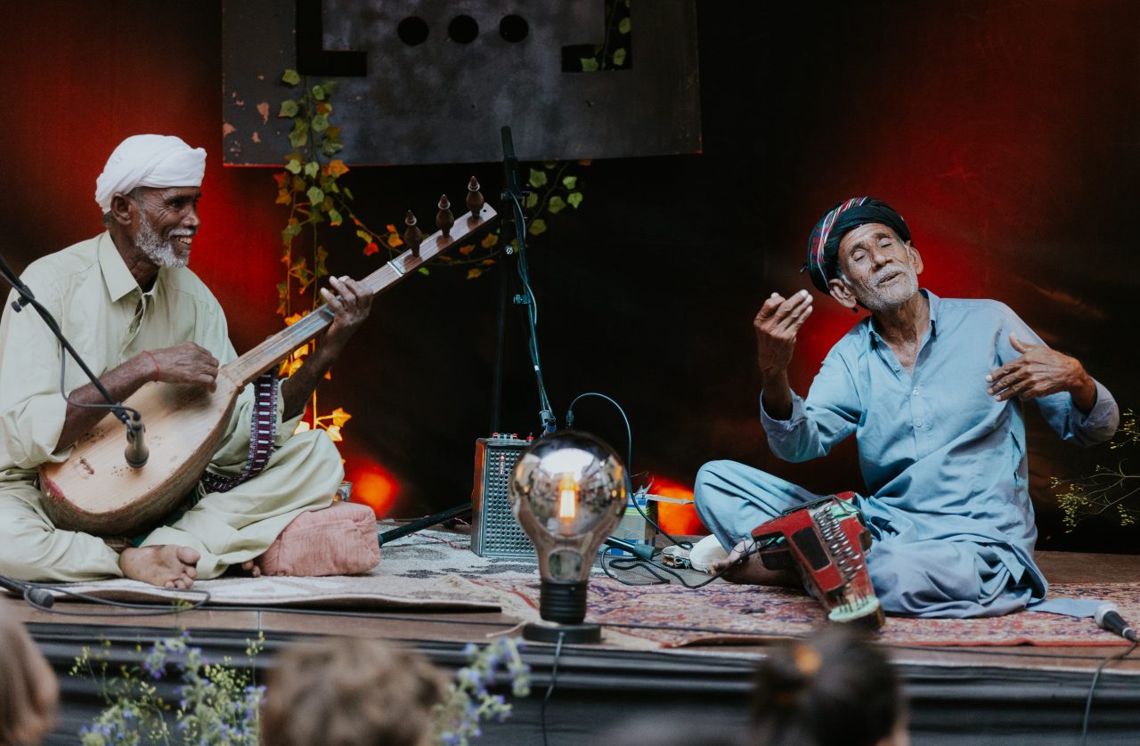 Two musicians play traditionally on stage, surrounded by soft light and a lively atmosphere.