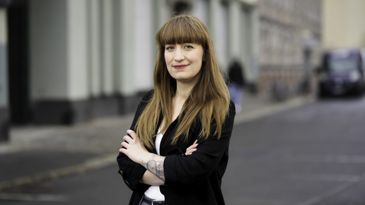Porträt einer lächelnden Frau mit langen, glatten Haaren, die in einer Stadt steht und die Arme verschränkt.