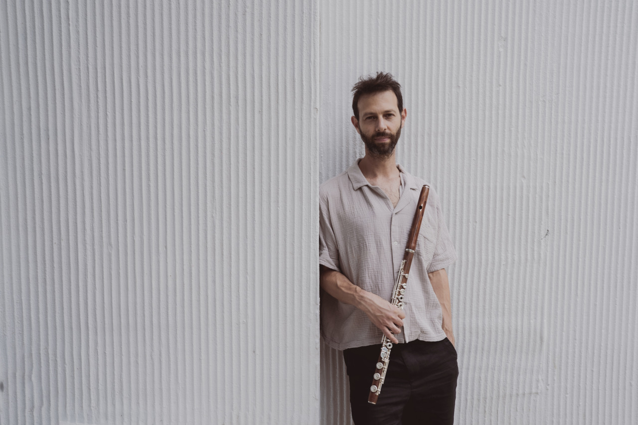 A man with a flute stands against a textured white wall, wearing a casual shirt and looking directly at the camera.