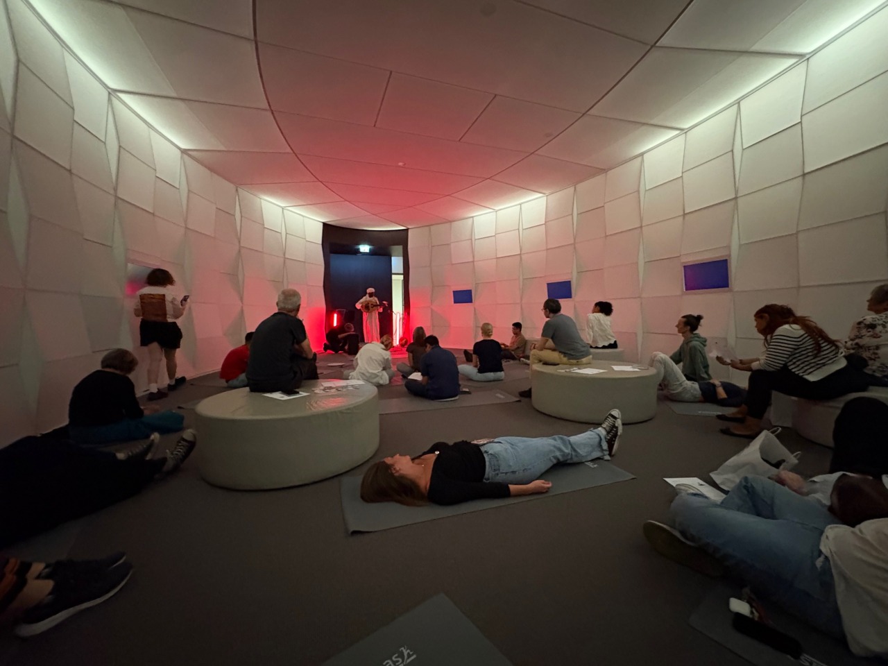 Eine Gruppe von Menschen sitzt und liegt in einem modernen, hellen Raum mit roten Lichtern und skulpturalen Wänden.