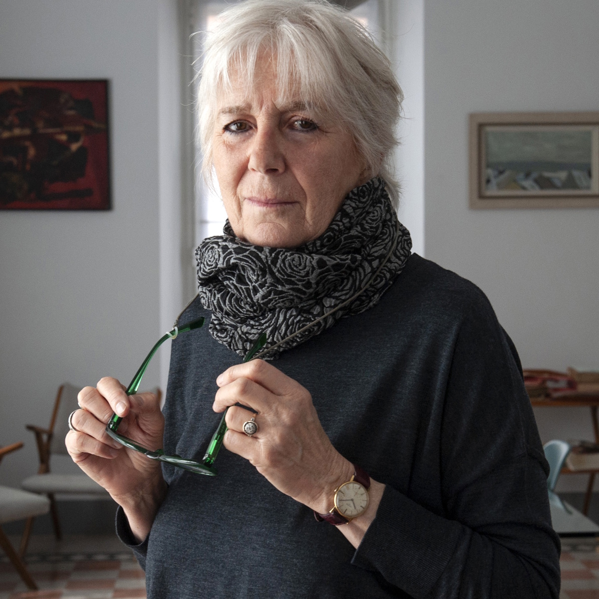 An older woman with gray hair, a scarf, and glasses looks directly at the camera while holding her glasses in her hand.