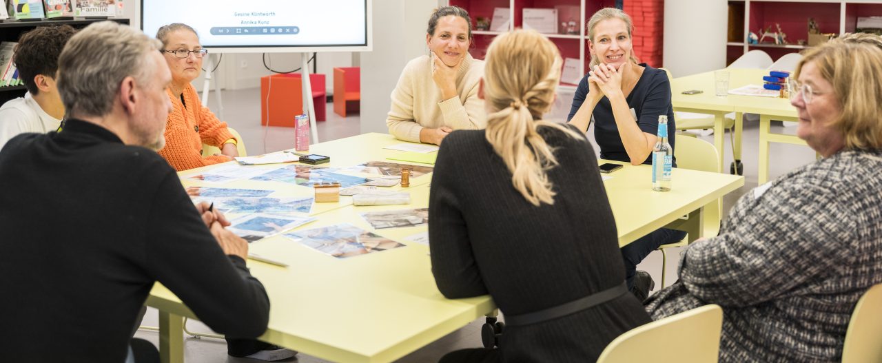 Eine Gruppe von Frauen und Männern diskutiert an einem Tisch, umgeben von Bildern und Notizen.